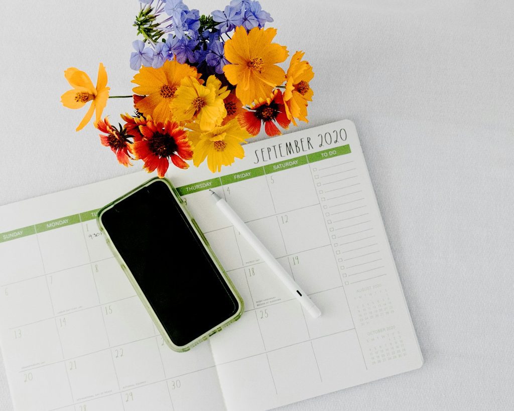 Telefono sobre una agenda blanca y una birome a un costado. De fondo unas flores de color amarillo. Ejemplificando el planner de google.