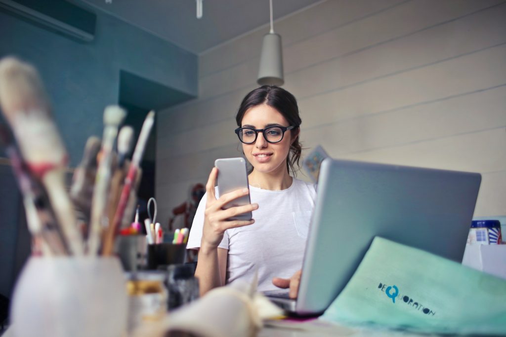 Mujer mirando el celular y con una notebook abierta delante de ella.